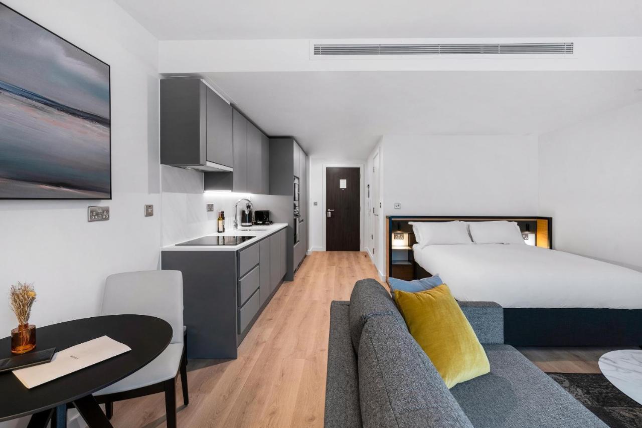 Citysuites 2 Aparthotel Salford  Exterior foto The photo shows a modern and stylish studio apartment. It features a kitchenette with dark cabinetry and a small dining table with two chairs. There's a cozy sofa with colorful cushions, positioned next to the table. In the background, a large bed wi
