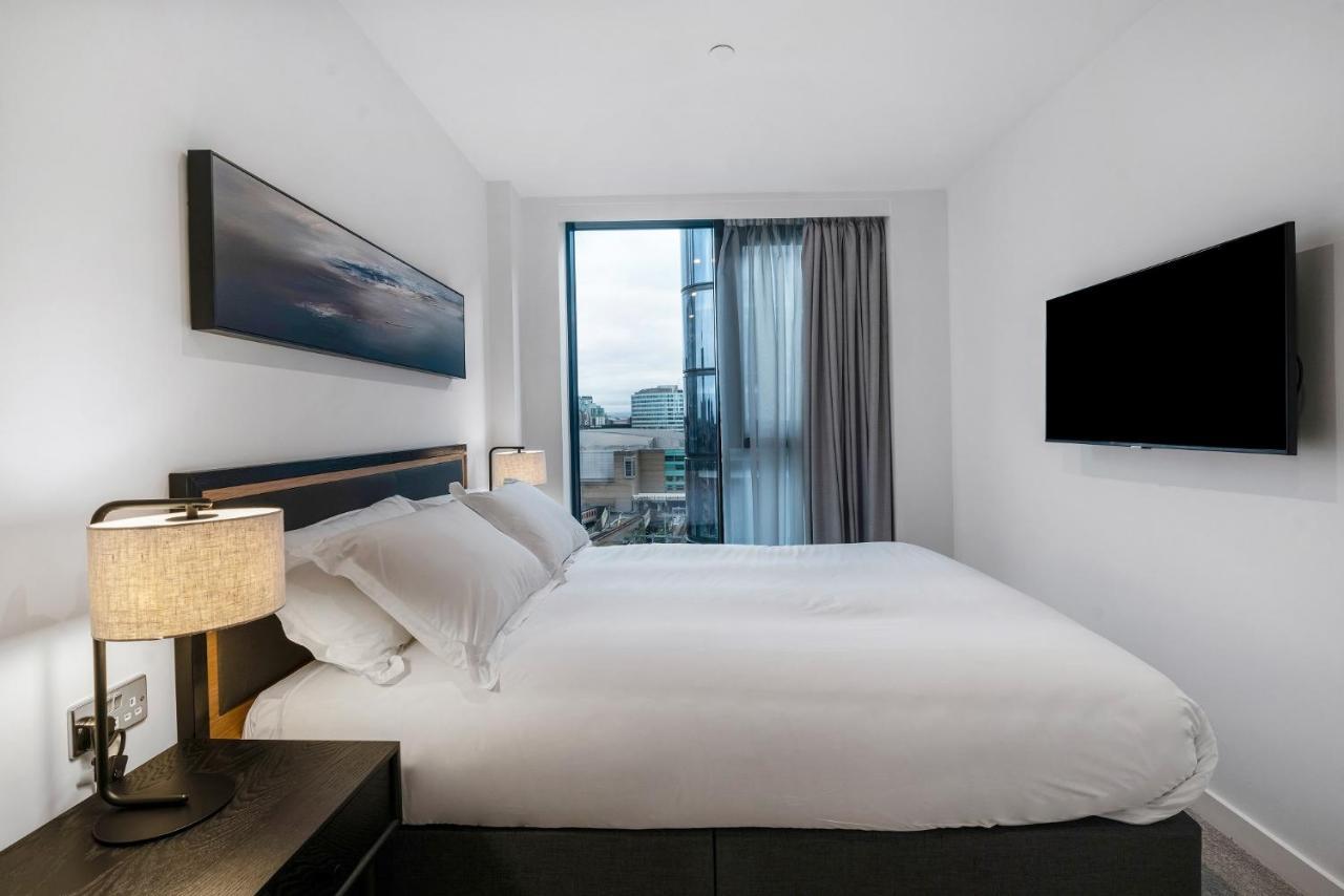 Citysuites 2 Aparthotel Salford  Exterior foto The photo shows a modern bedroom with a minimalist aesthetic. It features a large bed with white bedding and two pillows. On either side of the bed, there are bedside tables with lamps. A large window lets in natural light, covered by sheer curtains.