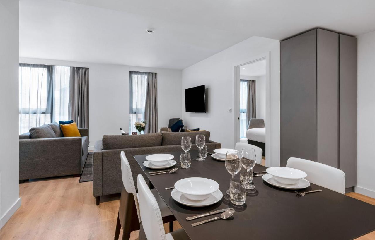 Citysuites 2 Aparthotel Salford  Exterior foto The photo shows a modern living space with an open layout. In the foreground, there is a dining table set for a meal, featuring multiple plates, glasses, and cutlery arranged neatly. In the background, there is a comfortable-looking gray sofa with a 