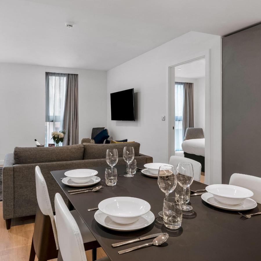 Citysuites 2 Aparthotel Salford  Exterior foto The photo shows a modern, well-lit living space that features a dining area and a sitting area. In the foreground, there is a dining table set for six, with white plates and crystal glasses arranged neatly. The table is surrounded by white chairs. 

