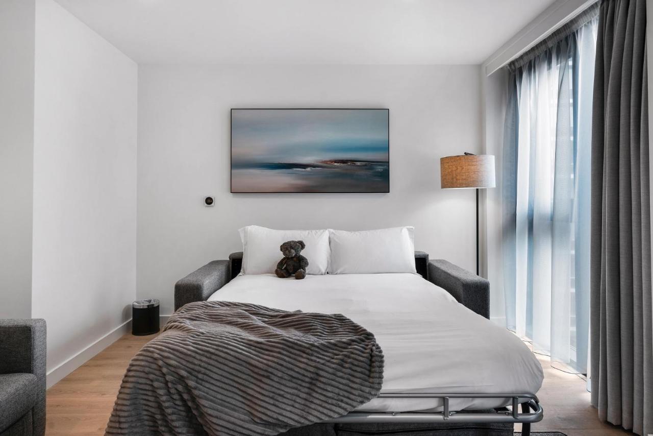 Citysuites 2 Aparthotel Salford  Exterior foto The photo shows a modern bedroom featuring a bed with white bedding and a decorative throw blanket. There is a teddy bear placed on the bed. A piece of abstract art is hanging on the wall above the bed. The room has a large window with sheer curtains