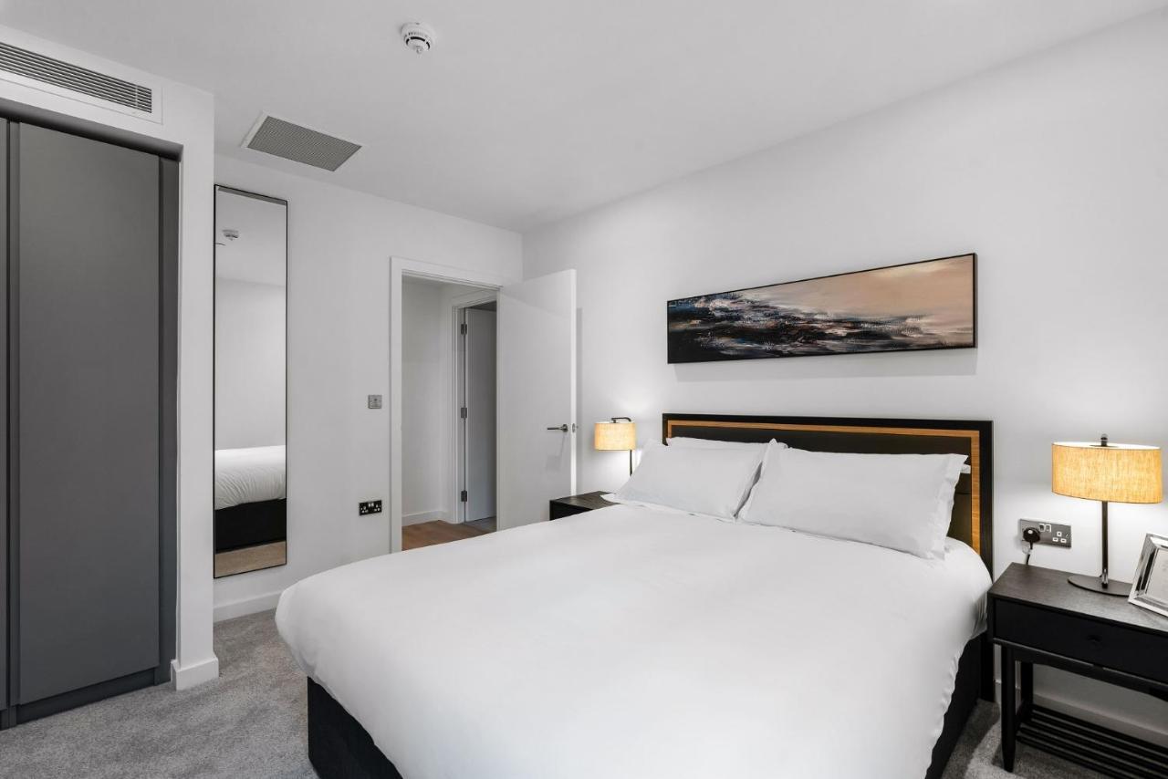 Citysuites 2 Aparthotel Salford  Exterior foto The photo shows a modern bedroom featuring a large double bed with white bedding. There are two bedside tables with lamps on either side of the bed. The walls are painted white, and there's a piece of artwork above the bed that depicts a landscape. A
