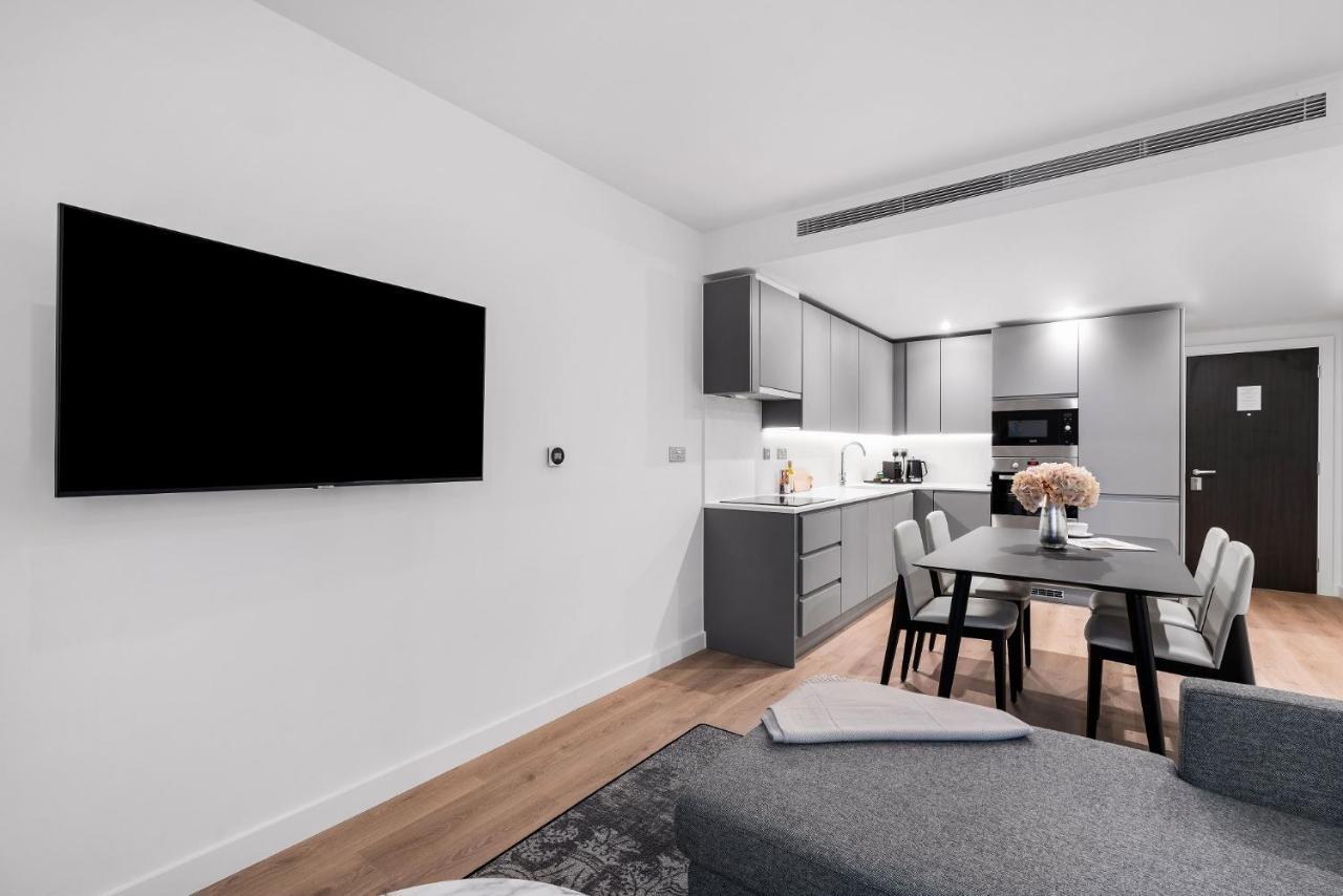 Citysuites 2 Aparthotel Salford  Exterior foto The photo shows a modern, minimalist living space. On the left, there is a large flat-screen TV mounted on the wall. To the right, a kitchen area features sleek gray cabinets and appliances, including a built-in microwave. In front of the kitchen, th