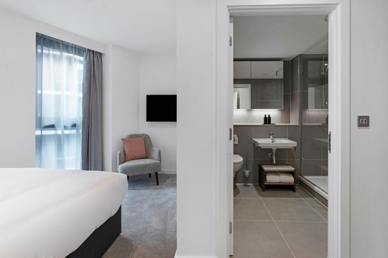 Citysuites 2 Aparthotel Salford  Exterior foto The photo shows a modern hotel room and bathroom setup. On the left, there is a bed with white bedding and a small sitting area that includes a gray chair and a pillow in a subtle pink shade. A flat-screen TV is mounted on the wall, providing enterta