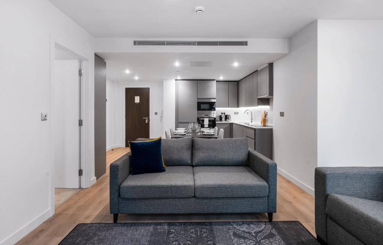 Citysuites 2 Aparthotel Salford  Exterior foto The photo shows a modern interior space, likely a living area or apartment. In the foreground, there is a gray sofa with a blue cushion positioned on a textured rug. To the back, there is an open kitchen with sleek cabinetry and appliances, visible a