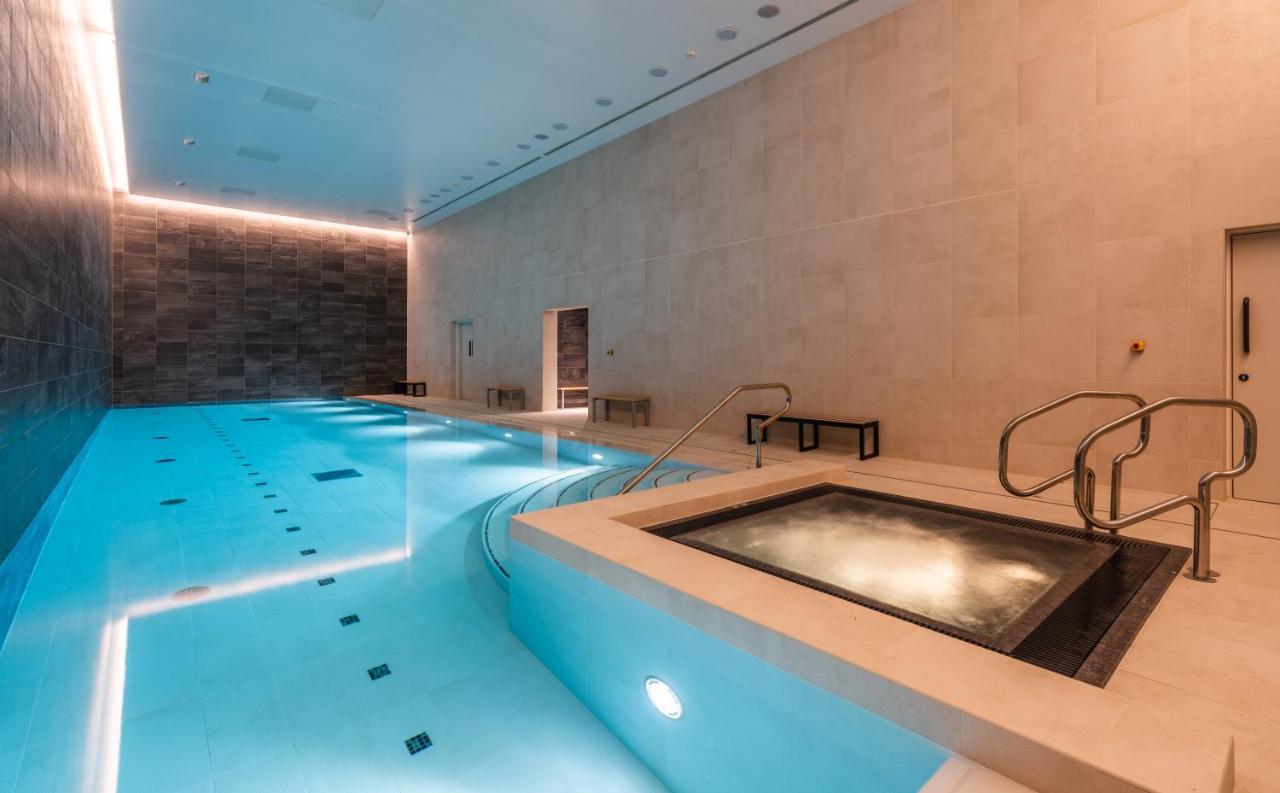 Citysuites 2 Aparthotel Salford  Exterior foto The photo shows an indoor swimming pool area. The pool is a bright blue color and features smooth, tiled surfaces. On one side, there is a hot tub area with steps leading into it. The walls are made of light-colored stone, and there is ambient lighti